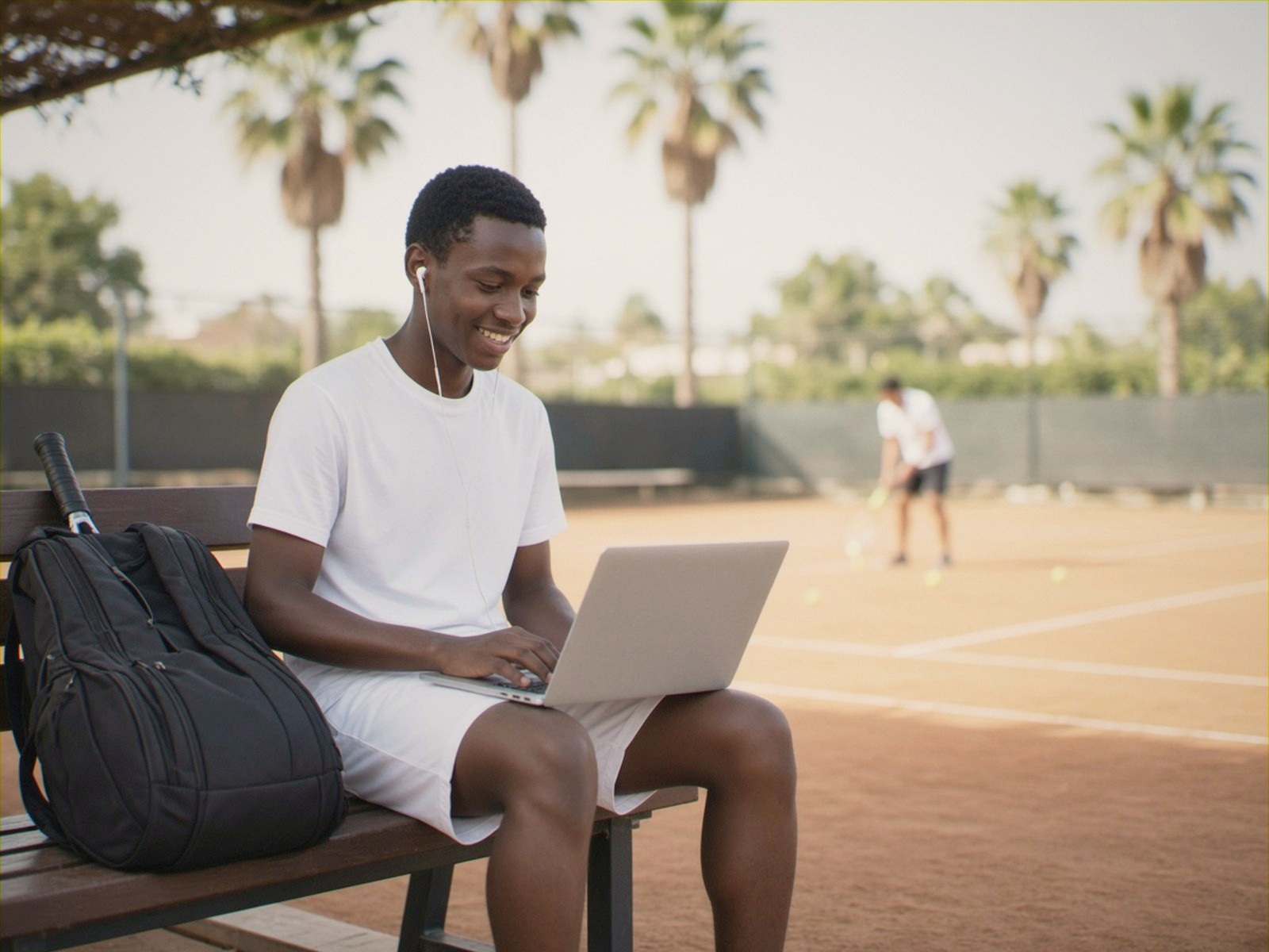 A teenage tennis player works on his laptop courtside at a training camp